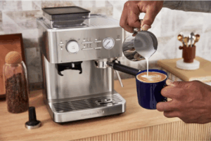 Un homme verse du lait chaud dans une tasse devant une machine à café KitchenAid en inox sur un plan de travail en bois.