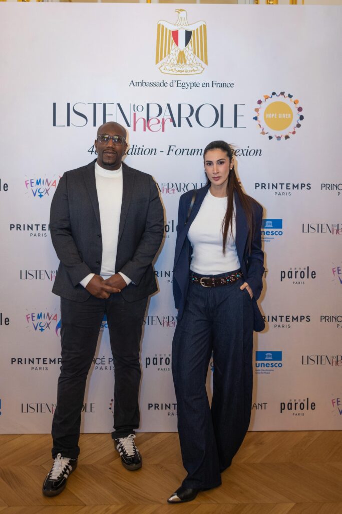 Lyna Mahyem avec un participant du Listen to Her Parole 2026 sur le fond d'un mur avec les logos des organisation participants de l'évènement