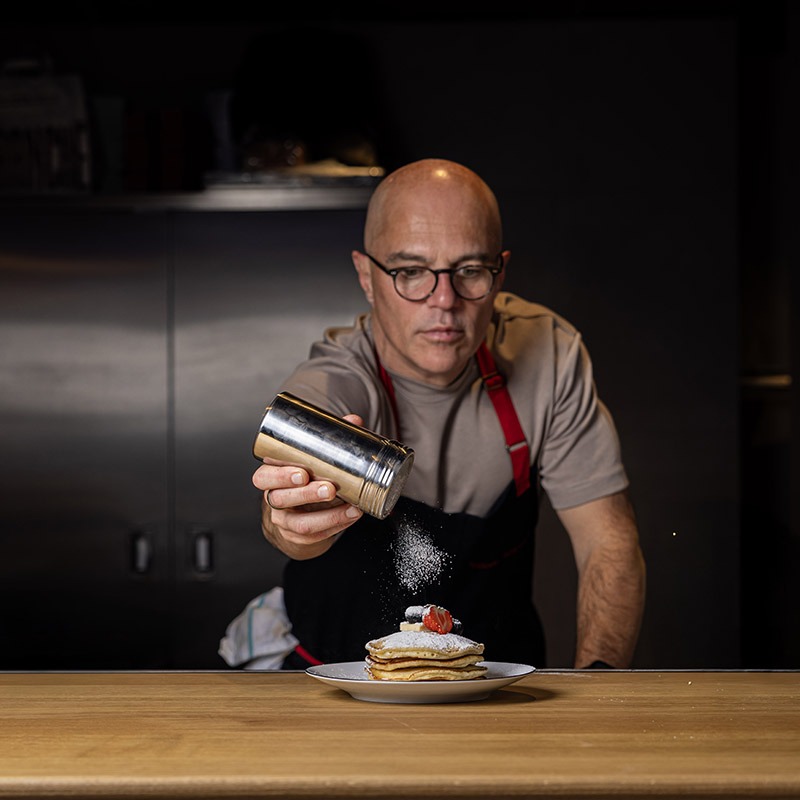 Chef Vittorio entrain de soupoudrer une pile de pancakes dans la cuisine du coffee shop