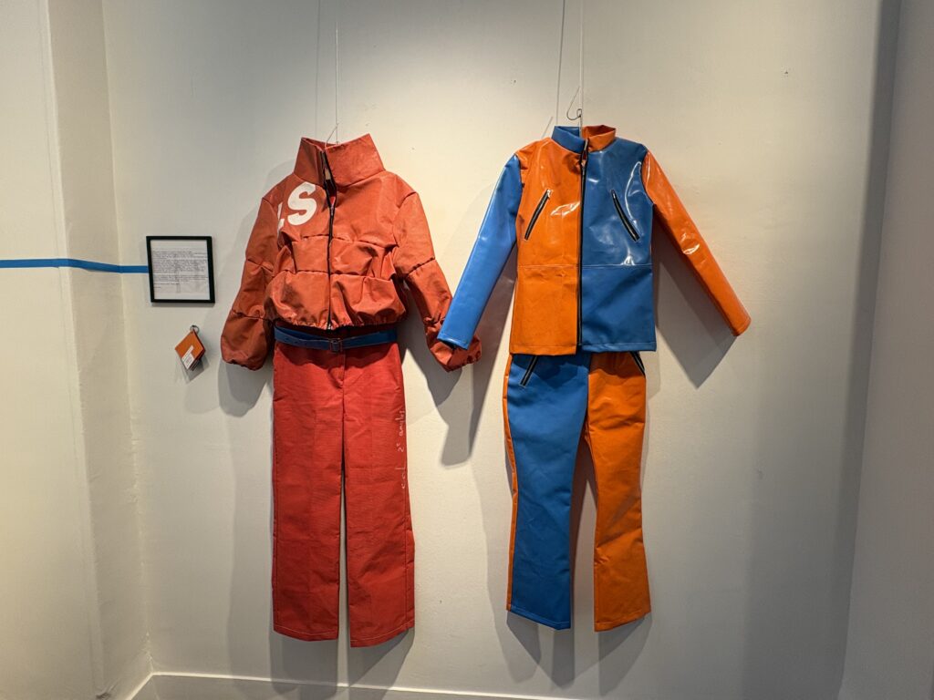 Exposition de la collection "Canon". À droite, le look 1 : un ensemble veste et pantalon en polyester recyclé orange et bleu. À gauche, le look 4 :un autre ensemble de sa collection : une doudoune orange avec un pantalon coupe évasé et une ceinture bleu. 